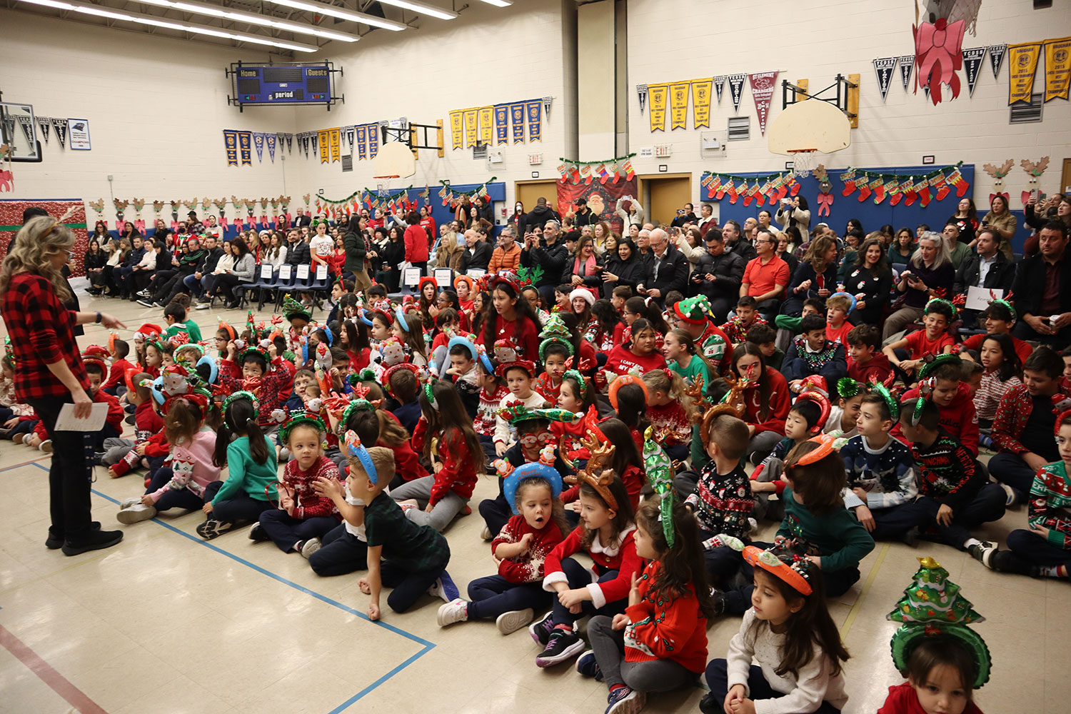 a large group of young students sitting on the floor of a school gym, wearing Christmas sweaters and headbands, with parents sitting behind the students on chairs.