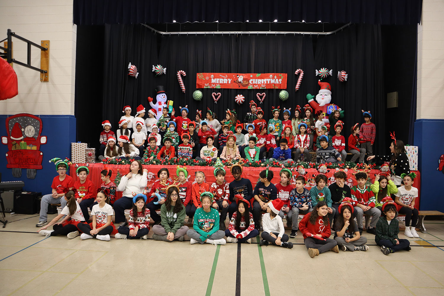  A large group of students in grades 4,5 and 6. sitting on the floor in front of a school stage as well as sitting on the stage. Everyone in the picture has a Christmas-themed headband on, and most are wearing a Christmas-themed shirt. 