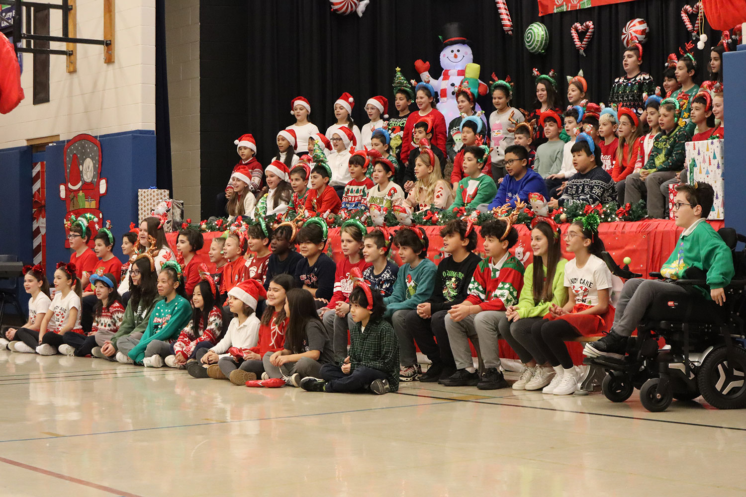 68 Grade 5 and 6 students wearing Christmas-themed headbands and shirts sitting on and in front of a decorated stage in a school gymnasium.