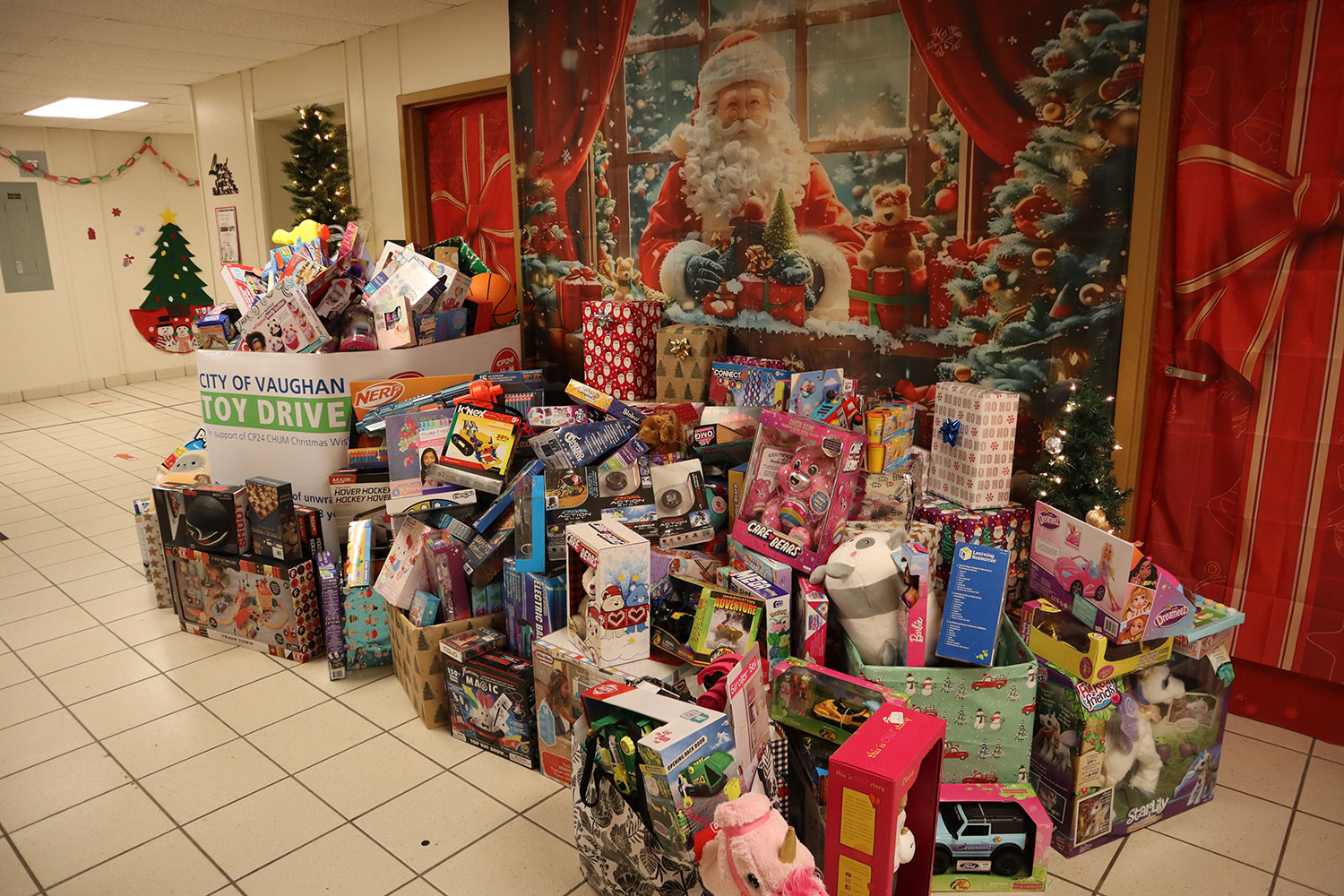 A large pile of unwrapped Christmas presents in a school hallway with a picture of Santa Claus on the wall behind the gifts.