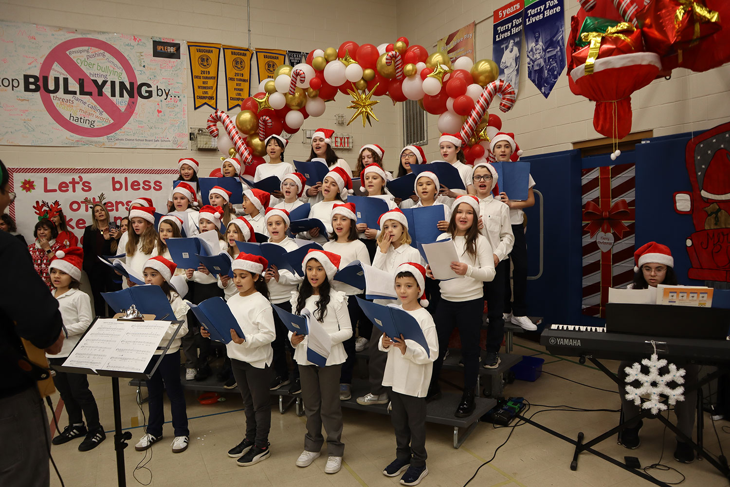 30 elementary school-age students dressed in white shirts and red Santa hats singing in a choir with a piano player to the side and a conductor in front of them.