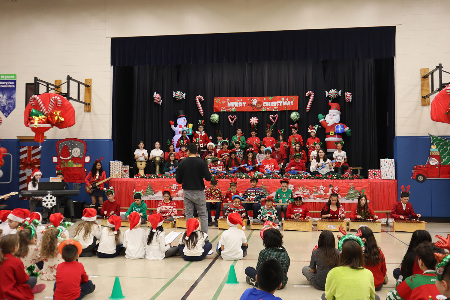 A group of students sitting on the stage and immediately in front of the stage playing xylophones and drums. A conductor is standing in front of them. They are inside a christmas themed decorated school gymnasium.