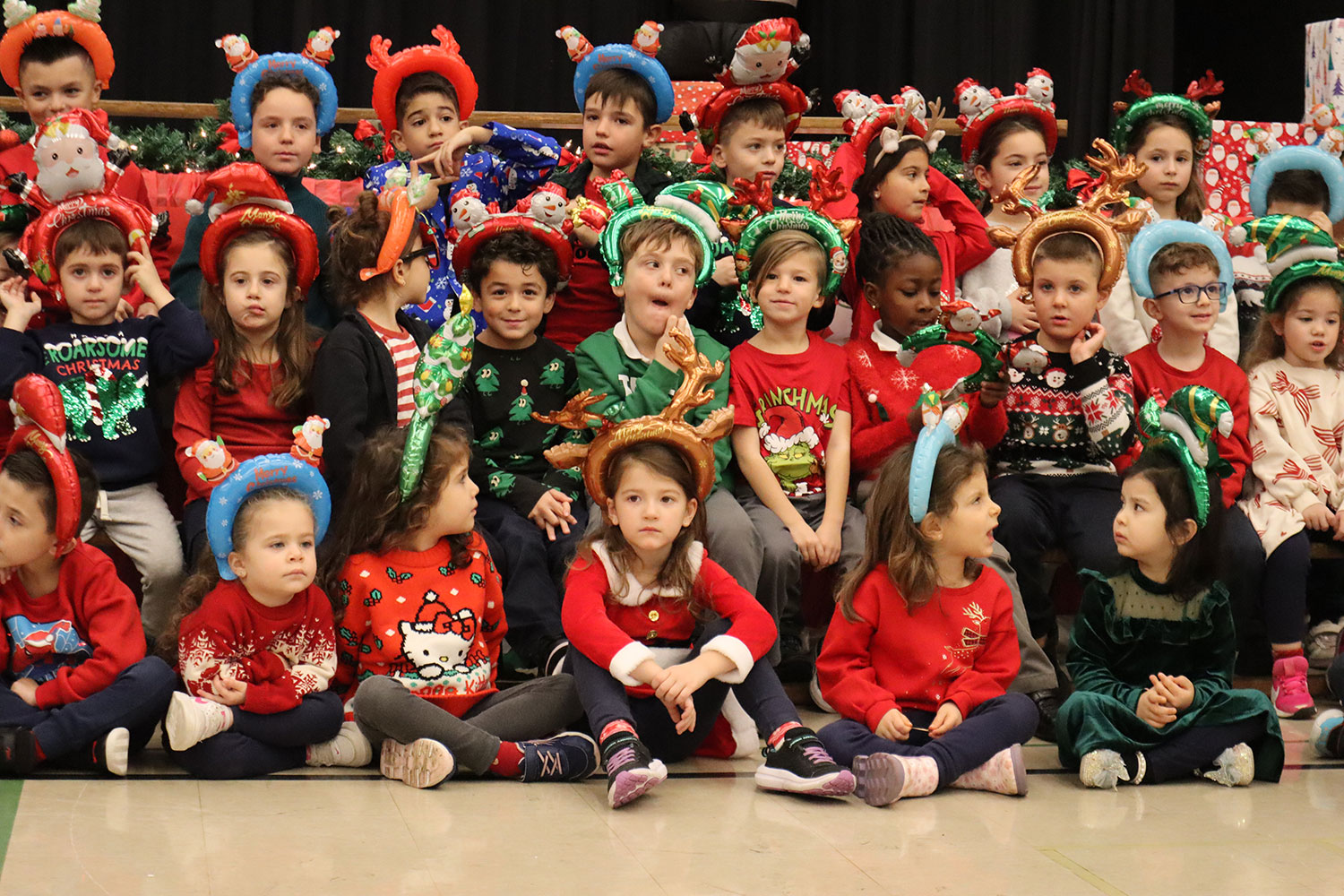 a group of kindergartner kids sitting on school benches wearing Christmas-themed outfits and headbands. 