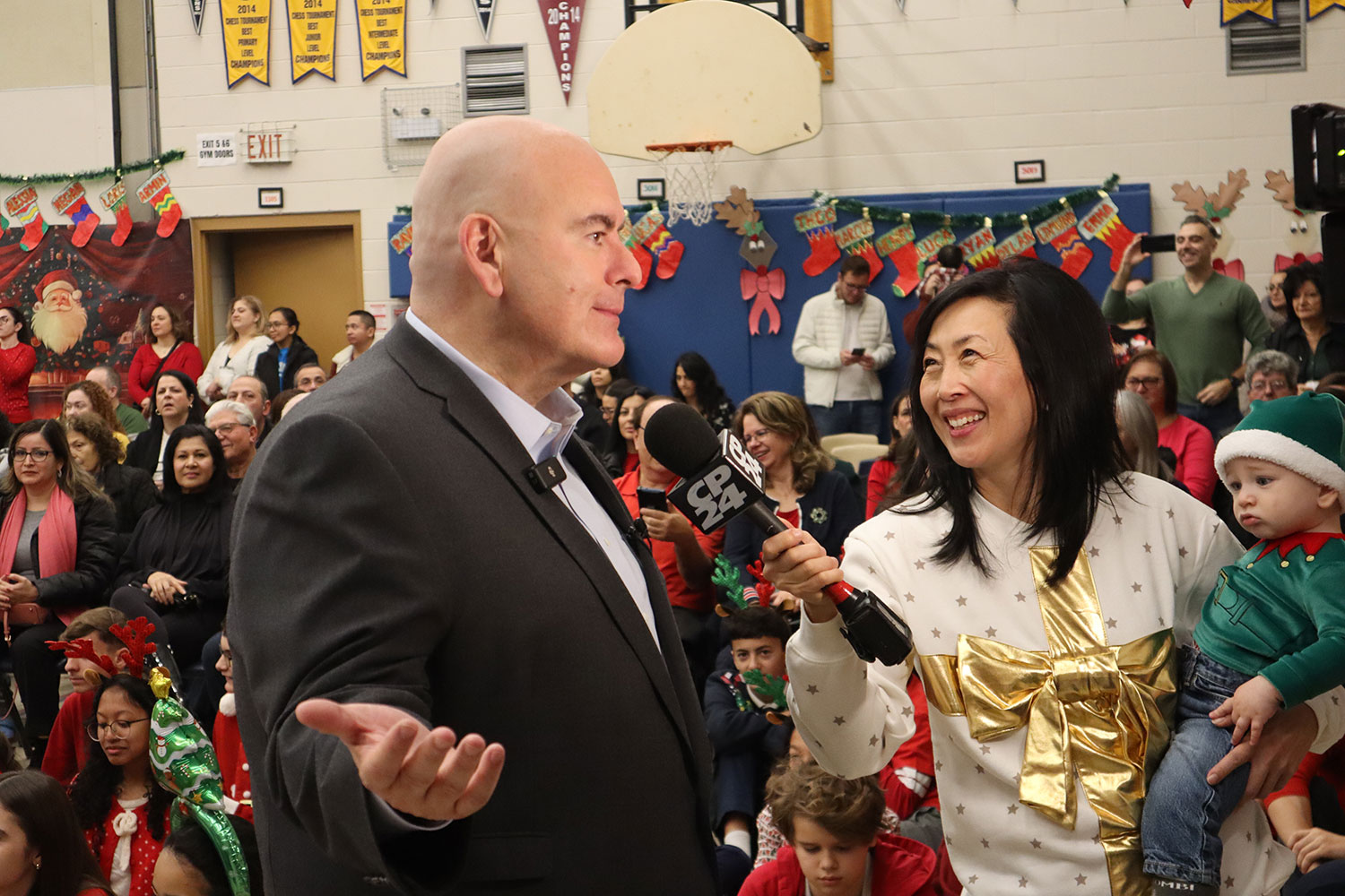 Vaughan Mayor Steven Del Duca being interviewed by a smiling journalist who is holding a baby standing in front of a group of parents who are sitting down and listening.