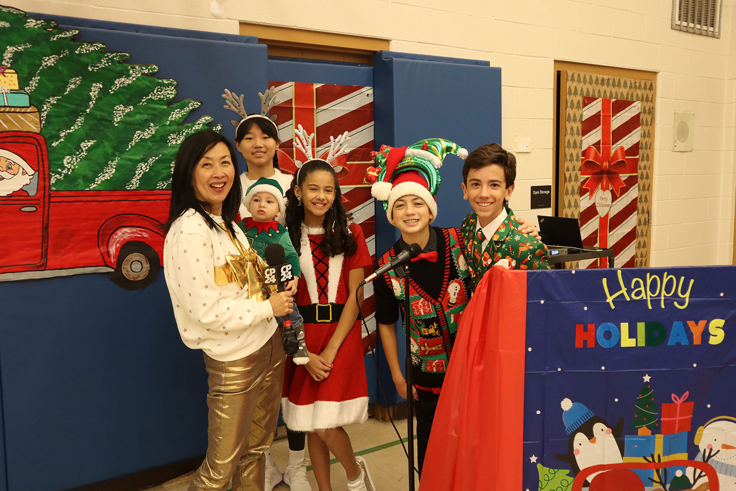 A smiling lady holding a baby and a microphone poses for a picture with four students in grades 7 and 8 who are wearing Christmas-themed outfits. 